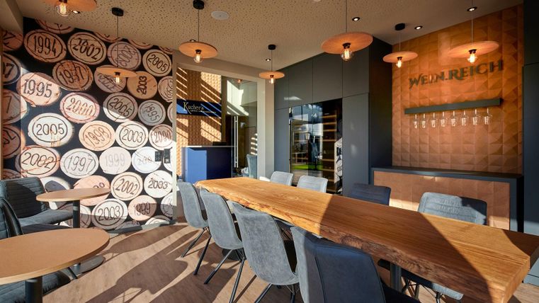 Een modern restaurant met een lange houten tafel en grijze stoelen. De muren zijn versierd met een stijlvol ontwerp en historische figuren.