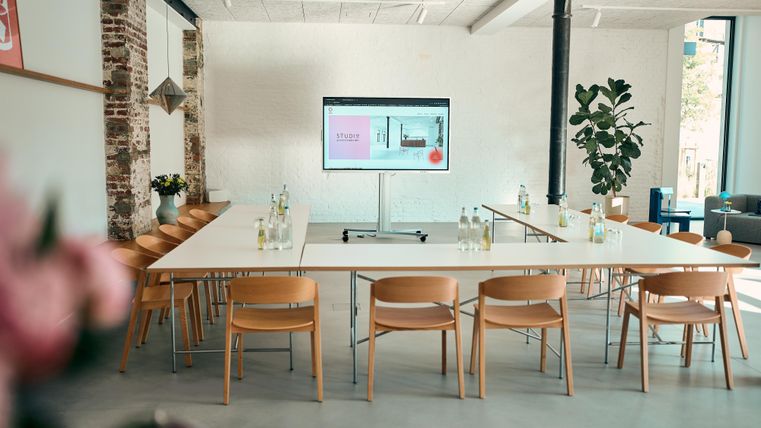 A modern conference room with a large table and chairs. A screen displays a presentation while plants create a pleasant atmosphere.
