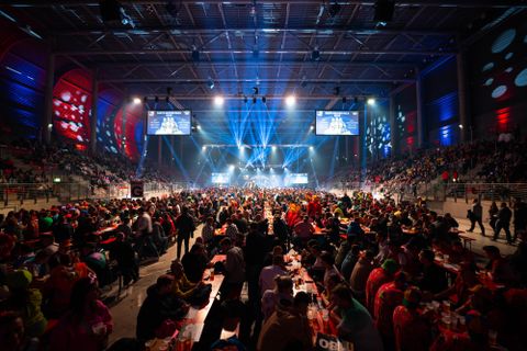 Ein großes Event mit einer vollen Halle. Viele Menschen sitzen an Tischen und genießen eine Live-Show mit bunten Lichtern.