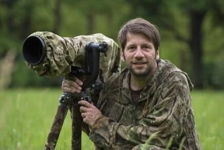 Ein Mann in camouflierter Kleidung steht in einer Wiese und hält ein Spektiv. Hinter ihm sind Bäume und eine grüne Landschaft sichtbar.