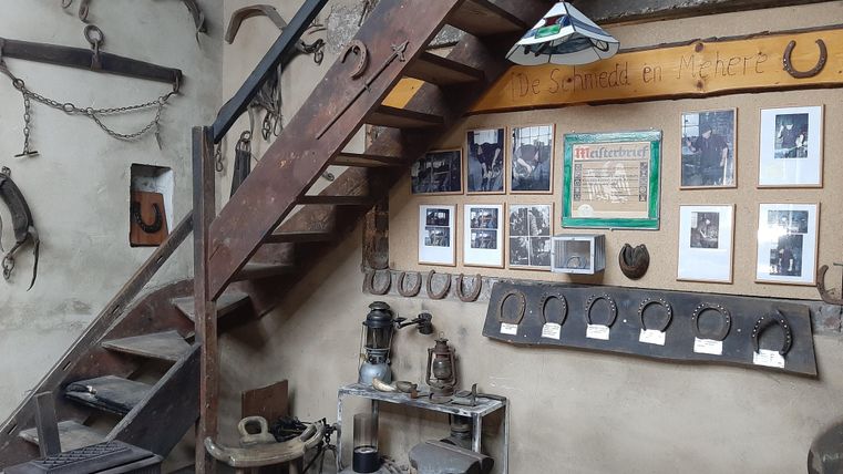 Eine alte Werkstatt mit einer Holztreppe und historischen Werkzeugen. An der Wand hängen Fotos und Hufeisen, die die Handwerkskunst dokumentieren.