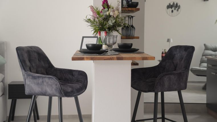 Een moderne eethoek met twee comfortabele, grijze krukken en een rustieke tafel. Op de tafel staan borden en een vaas met verse bloemen.