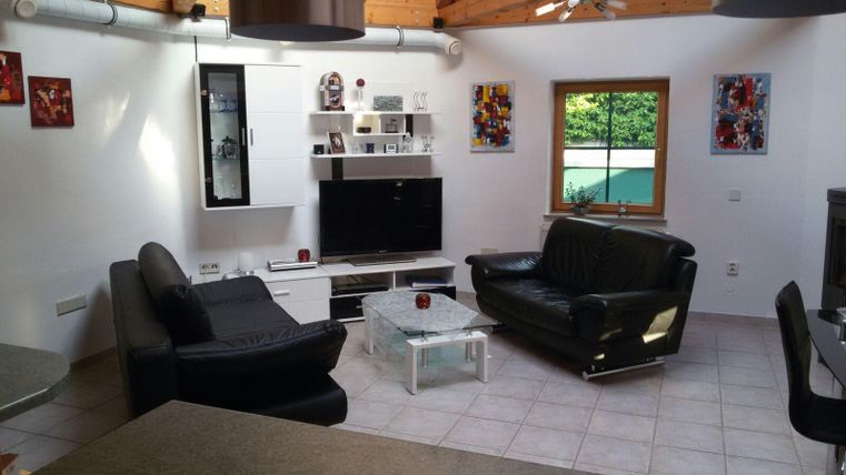 Een moderne woonkamer met zwarte banken en een televisie op een wit tv-meubel. Grote ramen laten veel daglicht binnen en de ruimte is stijlvol ingericht.