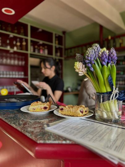 Deux assiettes de gâteau sont posées sur une table dans un café. En arrière-plan, une employée est visible, lisant un menu, tandis qu'un vase avec des hyacinthes se trouve sur la table.