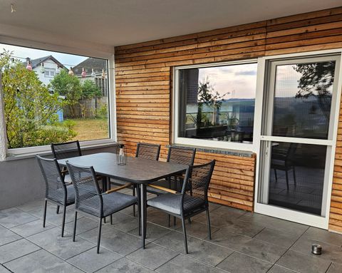 Een gezellige buitenruimte met een tafel en stoelen op een terras. Men ziet grote ramen en een groene omgeving.