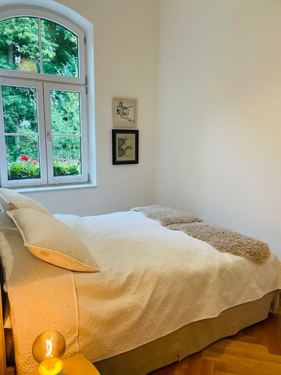 Une chambre confortable avec un grand lit et une lumière claire.
Par la fenêtre, on voit un beau jardin avec des plantes vertes.