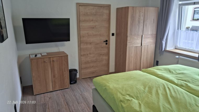 A modern bedroom with a double bed and light green blankets. On the wall, there is a television as well as a wooden wardrobe and a dresser.