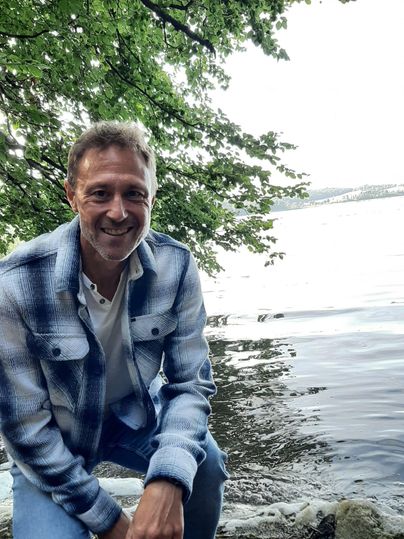 Ein Mann sitzt am Wasser und lächelt. Hinter ihm sind Bäume und eine ruhige Wasseroberfläche sichtbar.