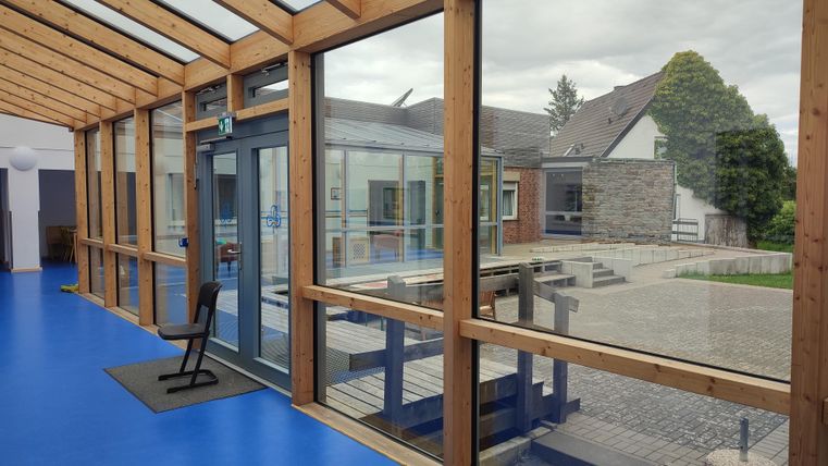 Une pièce lumineuse avec de grandes fenêtres et une vue sur une cour intérieure. Le sol est bleu et une chaise simple se trouve au premier plan.