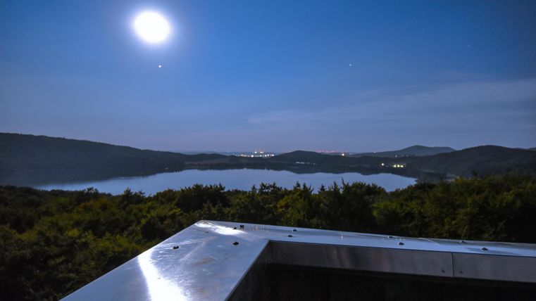 A serene night view with a clear moon over a tranquil lake. In the background, gentle hills and the soft light of the city can be seen.