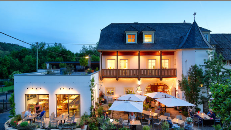 Un restaurant confortable avec terrasse et parasols. Il est crépuscule et le bâtiment émet une lumière chaleureuse.