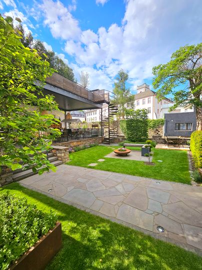 A beautiful garden with a stone area, lush greenery, and structured plants. In the background, a modern building and an outdoor staircase are visible.