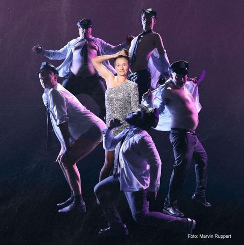 Eine Frau in einem glitzernden Kleid steht im Mittelpunkt, umgeben von Männern in Hemden und Mützen. Sie posieren schauspielerisch und zeigen verschiedene Posen.