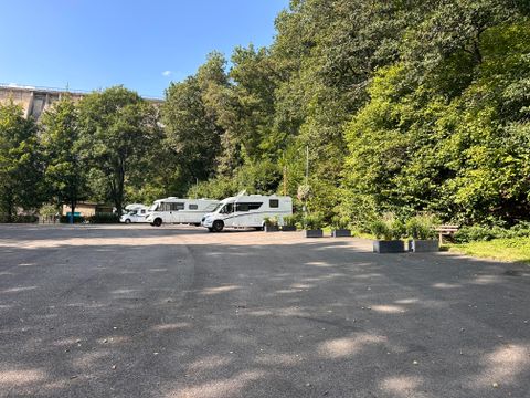 Ein ruhiger Parkplatz mit Wohnmobilen, umgeben von grünen Bäumen. Der Himmel ist klar und blau.