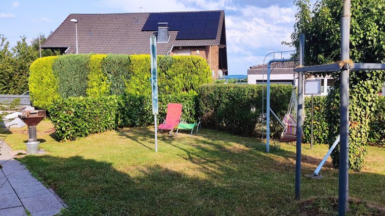 A well-kept garden with green grass, bushes, and some play equipment. In the background, a house and a partly cloudy sky can be seen.
