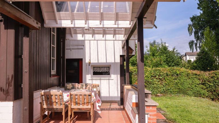 Une terrasse confortable avec des chaises et une table en bois, entourée d'un jardin verdoyant. En arrière-plan, un espace barbecue est visible.