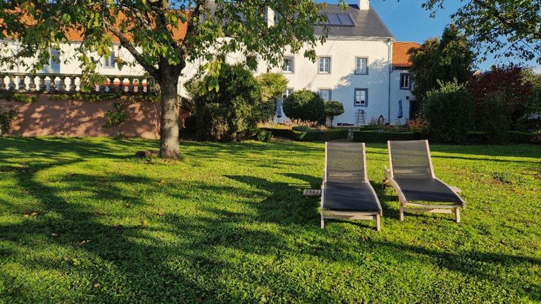 Two loungers on a green meadow in front of a white house. In the background, trees and bushes are visible.