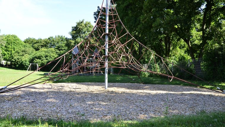 Een speelplaats met een klimrek van touwen staat op een weiland. Op de achtergrond zijn bomen en groen gras zichtbaar.