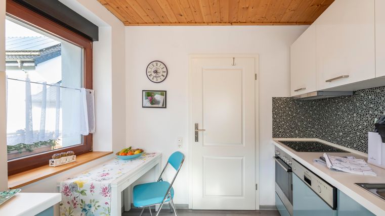 Een heldere keuken met moderne apparatuur en een houten plafond. Er is een kleine tafel met blauwe stoelen en een vensterbank met fruit.