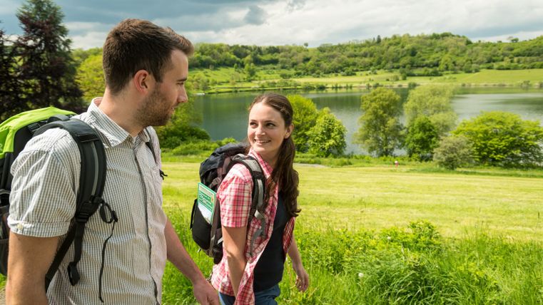 Deux randonneurs au Schalkenmehrener Maar avec des sacs à dos, en arrière-plan le lac et des collines vertes.
