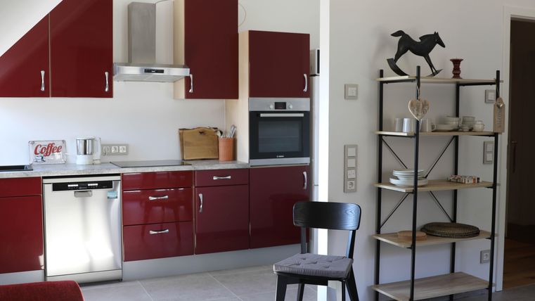 Eine moderne Küche mit roten Schränken und hellen Arbeitsflächen. Ein Regal mit Geschirr und Dekoration ergänzt den Raum.