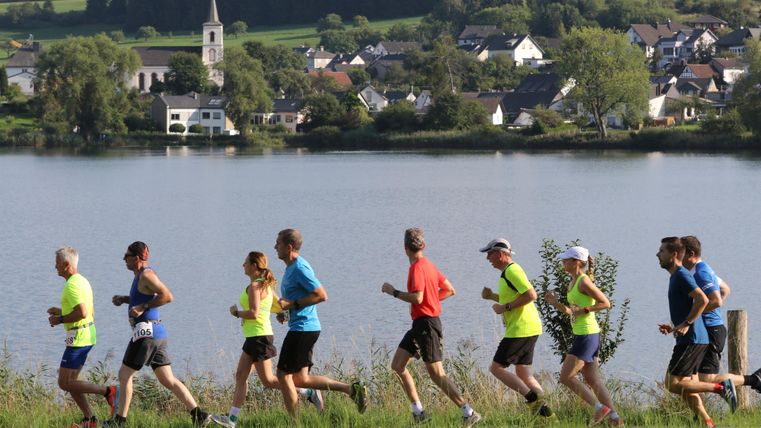 Eine Gruppe von Läufern joggt entlang eines Sees. Im Hintergrund sind grüne Wiesen und ein Dorf mit einer Kirche zu sehen.
