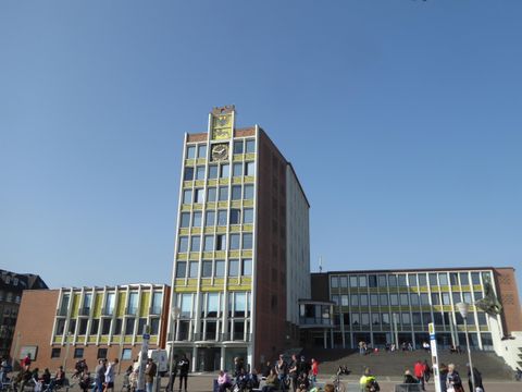 A modern building with a striking facade and a tall tower. People stand in front of it enjoying the sunny day.