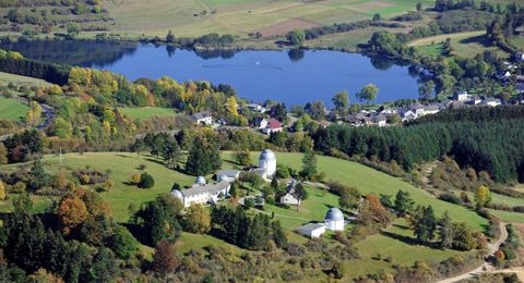 Eine ruhige Landschaft mit einem klaren See und bunten Bäumen. Im Hintergrund sind sanfte Hügel und kleine Gebäude zu sehen.