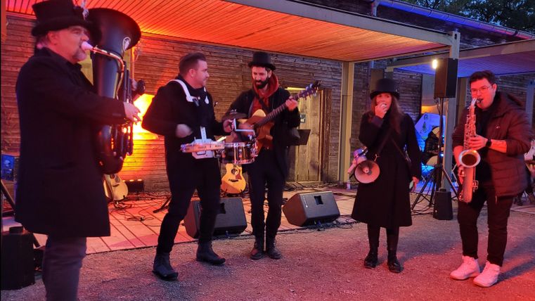 Un groupe joue de la musique en direct dans un coin extérieur convivial. Les artistes sont vêtus de noir et il y a des lumières colorées en arrière-plan.