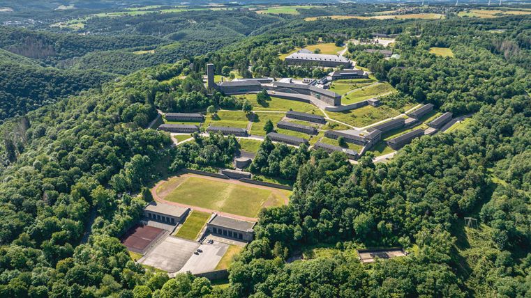 An aerial shot of historical buildings surrounded by dense forests and gentle hills. The sky is clear, and there are some wind turbines in the background.
