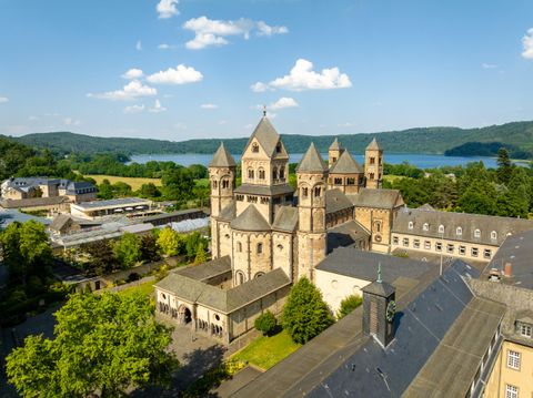 Ein beeindruckendes Klostergebäude mit Türmen, umgeben von Bäumen und Wiesen. Im Hintergrund erstreckt sich ein ruhiger See unter einem blauen Himmel.