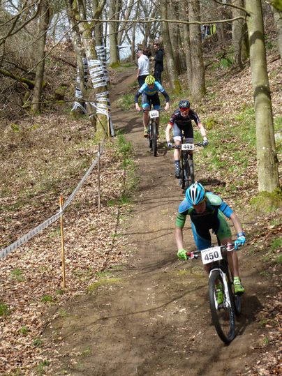 Meerdere mountainbikers rijden over een smal pad door een bos. Het parcours is gemarkeerd met afzetlinten en het is zonnig.