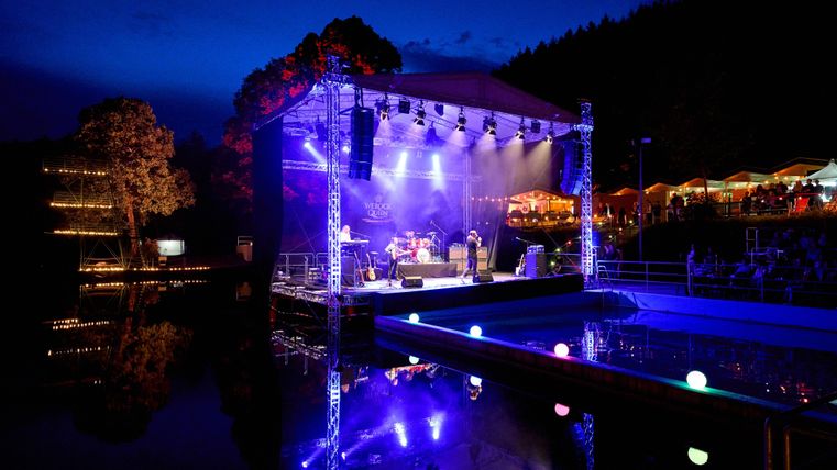 Een verlichte scène met muzikanten staat aan de oever van een water. Op de achtergrond zijn feestelijk versierde kramen te zien.