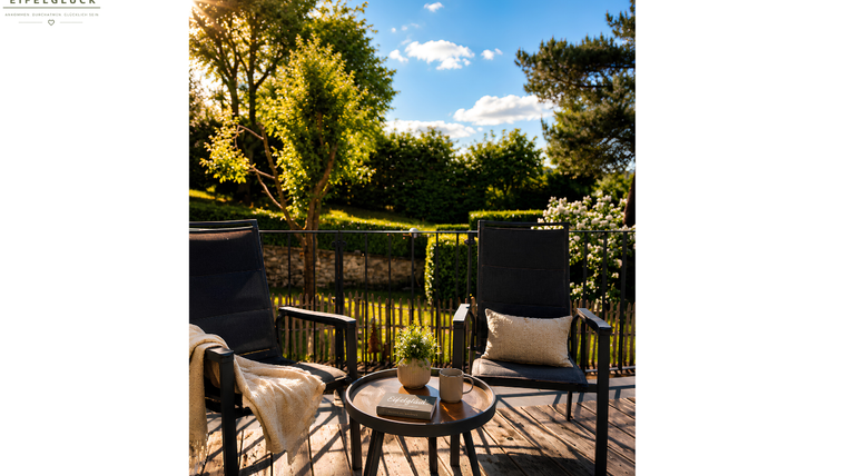 Gemütliche Sitzecke auf einer Terrasse mit Blick ins Grüne. Sonniges Wetter und blauer Himmel schaffen eine einladende Atmosphäre.