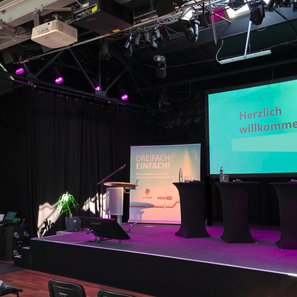 Een podium met een podium en twee statafels. Op de achtergrond bevindt zich een scherm met de tekst "Hartelijk welkom".