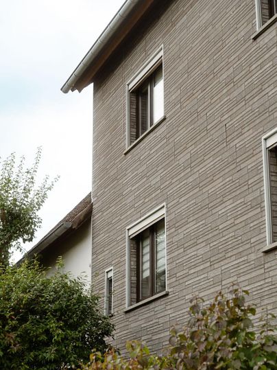 Une vue moderne d'un immeuble résidentiel avec une façade grise et plusieurs fenêtres. Des plantes vertes entourent le bâtiment et donnent à la scène une ambiance conviviale.
