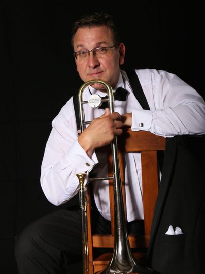 Ein Mann in formeller Kleidung sitzt auf einem Stuhl und hält eine Posaune. Der Hintergrund ist schwarz, was den Fokus auf den Musiker und sein Instrument legt.