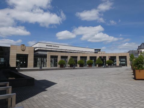 Ein modernes Gebäude mit großen Fenstern und einem Platz davor. Der Himmel ist klar mit einigen Wolken.