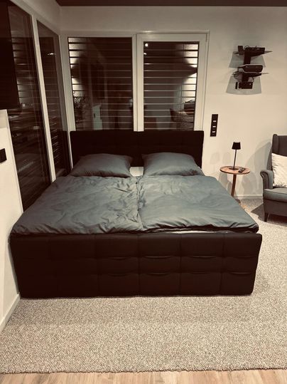 Une chambre moderne avec un grand lit double et des draps gris. À côté du lit se trouve une petite table et un fauteuil.