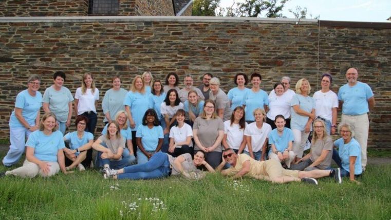 Eine Gruppe von Menschen posiert fröhlich vor einer Steinmauer. Sie tragen helle Kleidung und sind in einem grünen Bereich zusammen versammelt.