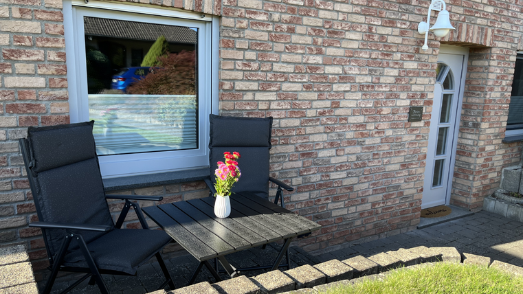 Une terrasse confortable avec deux chaises noires et une table en bois. Sur la table se trouve un vase avec des fleurs colorées devant une maison en brique.
