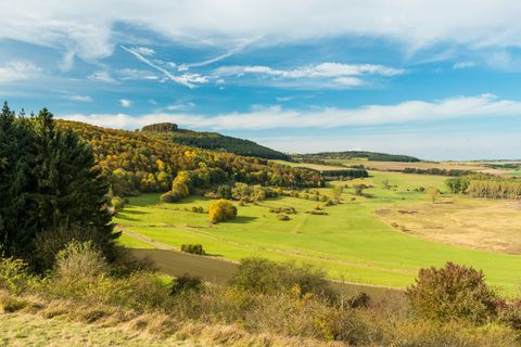 Landschaft mit grünen Wiesen, bewaldeten Hügeln und blauem Himmel.