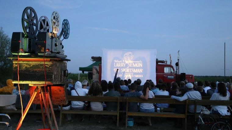 Ein Freiluftkino mit Zuschauerinnen und Zuschauern, die auf Holzbänken sitzen. Im Vordergrund ist ein alten Filmprojektor zu sehen, der einen Film auf eine Leinwand projiziert.