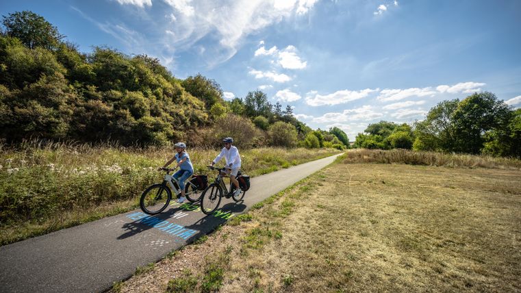 Deux cyclistes roulent sur un chemin calme à travers la nature. En arrière-plan, on peut voir des arbres verts et un ciel bleu.