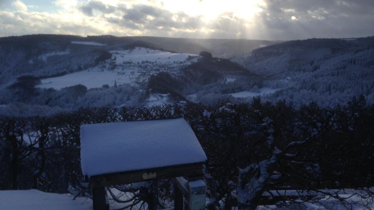 A winter landscape with snow-covered hills and trees. In the background, sun rays shine through the clouds.