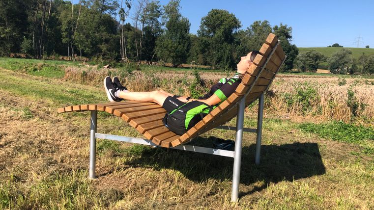 Persoon ligt op een gebogen houten bank in een landelijke omgeving.