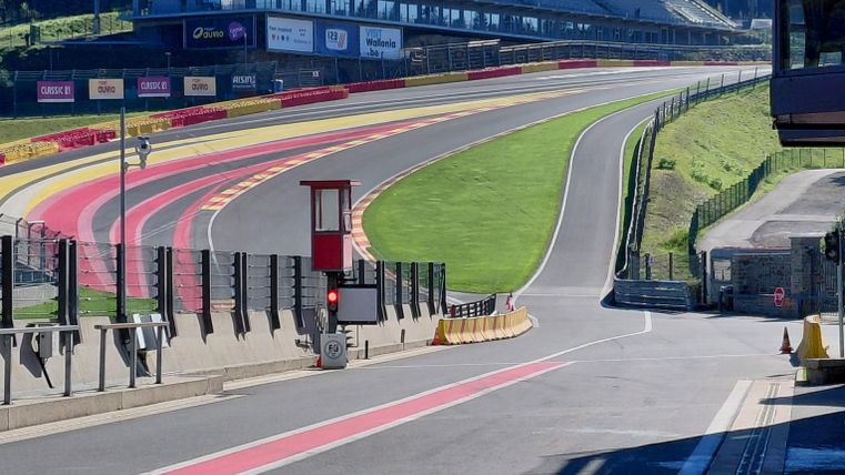 Une piste de course avec un long virage et une vue sur les tribunes. À l'arrière-plan, des arbres et un ciel dégagé sont visibles.