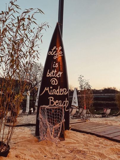 Een bord in de vorm van een surfplank toont de boodschap: „Life is better @ Minden Beach“. De achtergrond heeft een ontspannen strandatmosfeer met zand en planten.