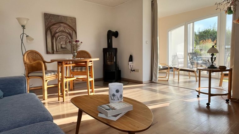 Ein helles, einladendes Wohnzimmer mit einem blauen Sofa und einem Holztisch. Große Fenster lassen viel Tageslicht herein und bieten einen Blick auf den Außenbereich.