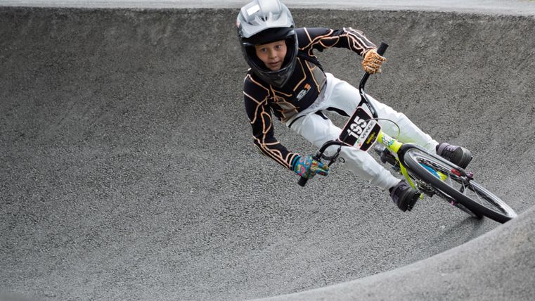 Un jeune rider de BMX roule à haute vitesse sur un parcours sinueux. Il porte un casque et des vêtements de cyclisme spéciaux.